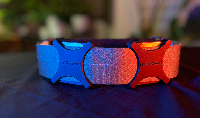 A wearable wellness device with blue and red sections, branded as HYDRAWAV3, displayed on a dark surface under blue and red lighting.